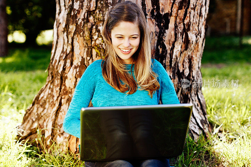 微笑的年轻美女坐在树下，忙着她的笔记本电脑
