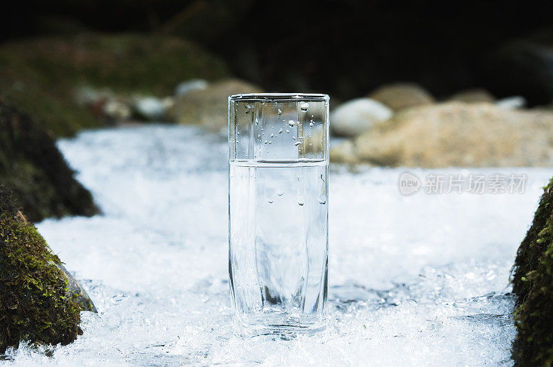 透明的玻璃玻璃与饮用的山水在冬天站在冰壳上，背景是一个干净的，结霜的河岸。饮用山泉水的概念和生产生态清洁的矿泉饮水