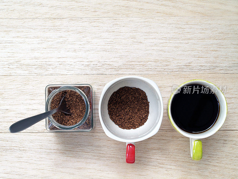 速溶咖啡用银勺子装在透明的小玻璃罐子里，咖啡粉装在杯子里，另外一个杯子里是在木地板上煮黑咖啡