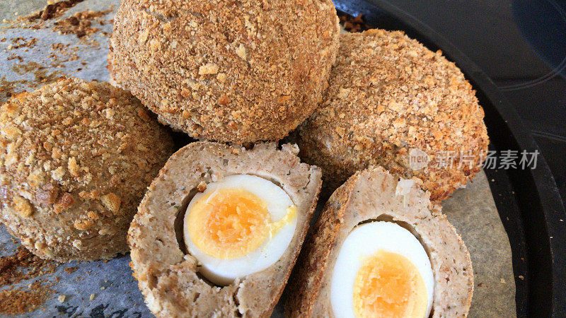 自制健康苏格兰鸡蛋切成两半，显示煮熟的蛋清和橘黄色的蛋黄，煮熟的鸡蛋包裹在香肠肉和棕色全麦面包屑，在烤箱中烘烤，作为健康的饮食零食，而不是不健康的油炸