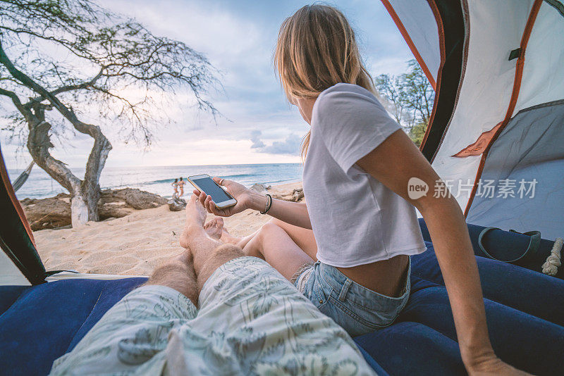 年轻夫妇在帐篷里用手机，两人在沙滩上露营