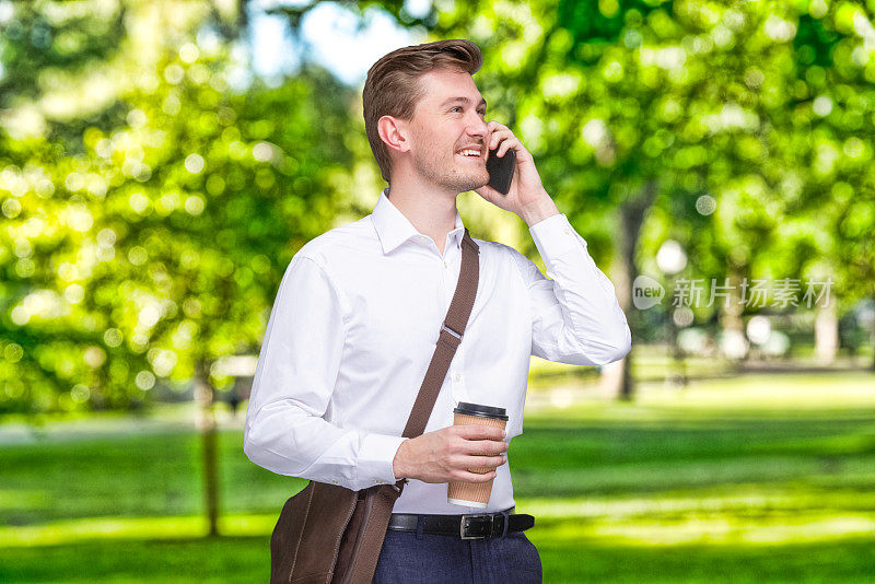 白人男性经理站在公园前，带着肩包，拿着包，使用智能手机