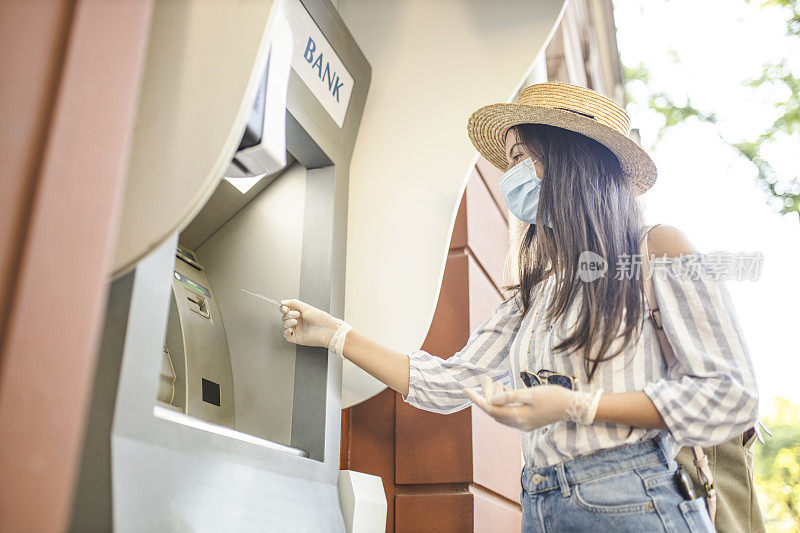 戴着防护口罩和外科手套的妇女站在城市街道上，使用自动取款机取钱。冠状病毒或Covid-19病毒大流行概念。