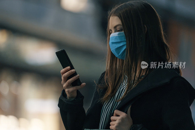 2019冠状病毒病期间，年轻女孩佩戴口罩在城市商业区夜间使用智能手机