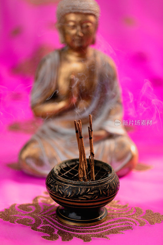 在一个没有聚焦的金佛前面的香碗中冒烟的香和一个美丽的刺绣的粉红色印度教纱丽绳的特写