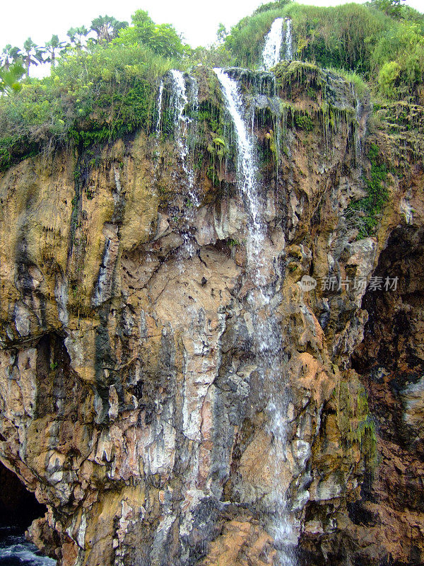 一条小瀑布从石崖上泻下。悬崖顶上有绿色的灌木和树木
