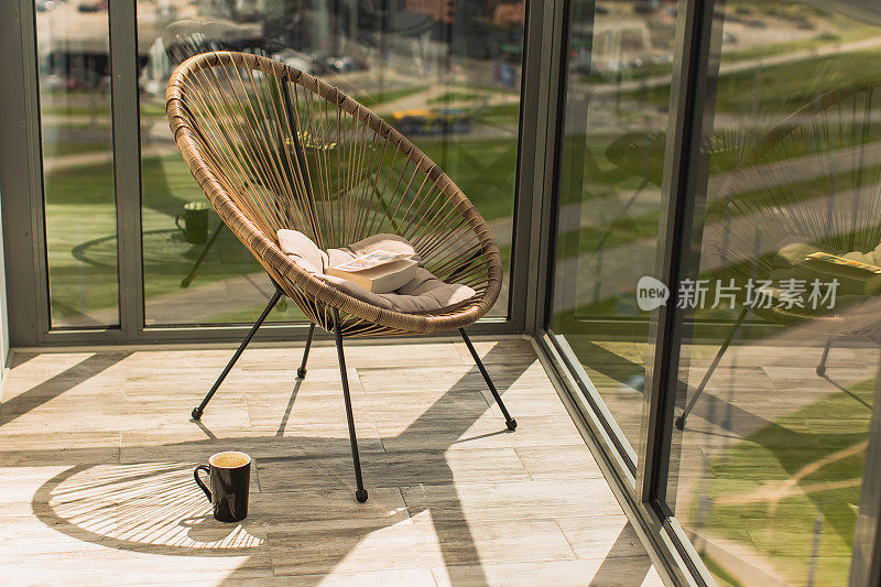 阳台上有靠垫的柳条椅。窗外的景色很美。出租的房子。上午休息。休闲的概念。阳光明媚的一天。地板上有一杯茶。书在椅子上。在家教育。