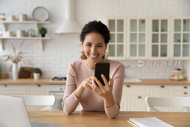 兴奋的女人拿着智能手机，在现代化的厨房里在线聊天