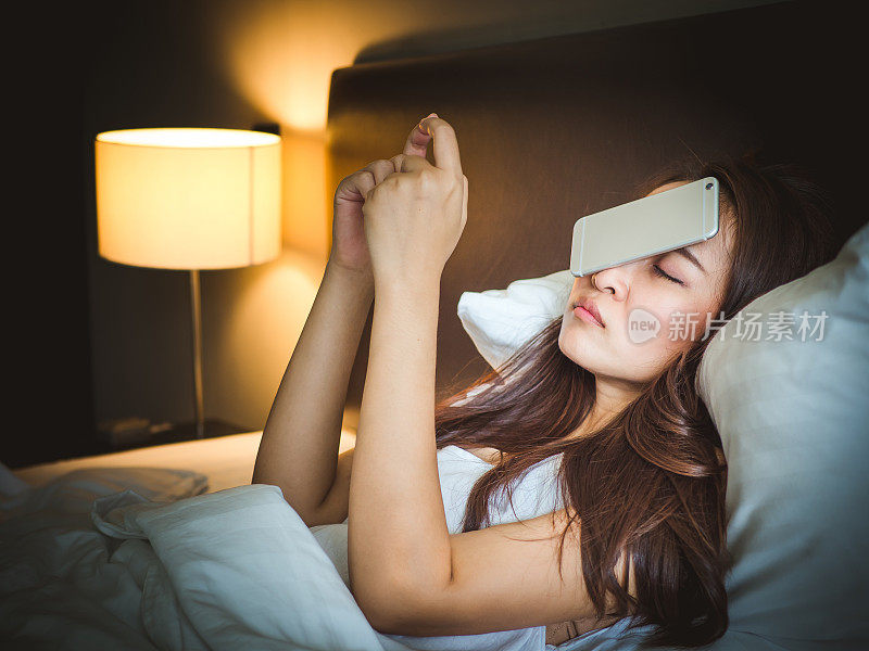 睡在白色床上的亚洲女人，把智能手机掉在脸上