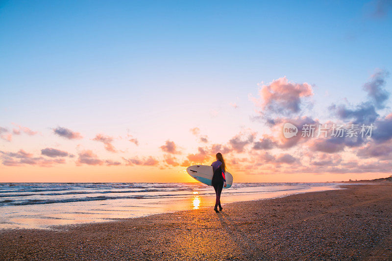 冲浪妇女与冲浪板在海滩上日落或日出，海洋和波浪与温暖的颜色
