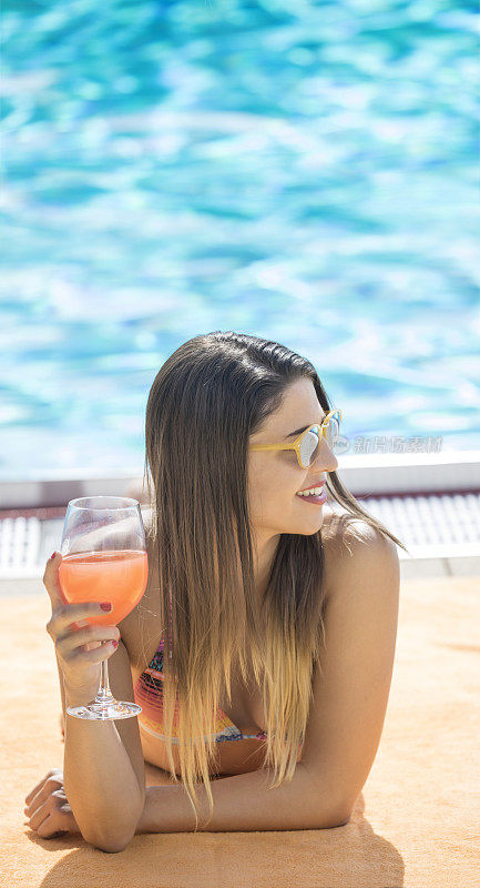 美丽的年轻女子日光浴在比基尼附近的游泳池喝鸡尾酒