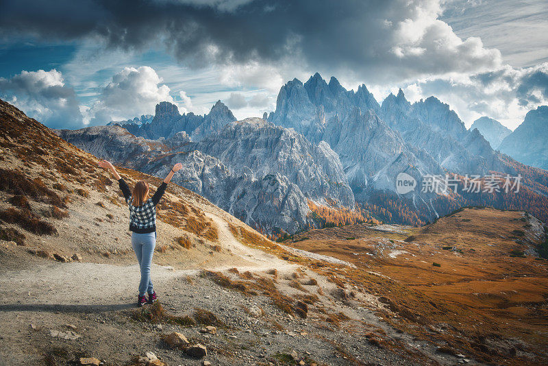在意大利多洛米特的秋天日落时，年轻的女子举起手臂，站在雄伟的山峰上。风景与快乐的女孩，戏剧性的天空与云，橘子树，高岩石在意大利阿尔卑斯山在秋天