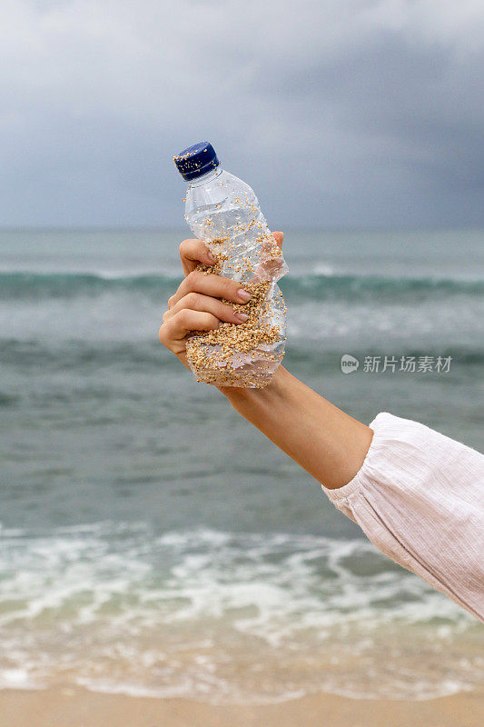 蓝天背景下，手里拿着一个塑料瓶。从海滩上收集垃圾的行动。Trash-free海域概念。一次性塑料是人类的嗜好，它正在破坏我们的计划