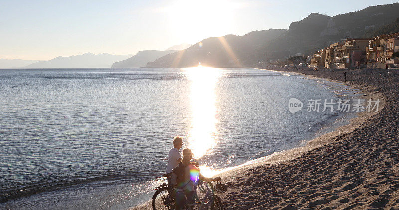 一对夫妇在日落时分沿着海滩海岸线推着自行车