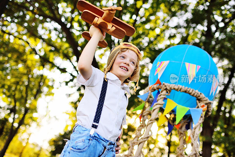小男孩穿着白色衬衫，背带，卷发，戴着飞行员的帽子和眼镜，在一个篮子和一个蓝色气球的背景下玩着一架木制飞机
