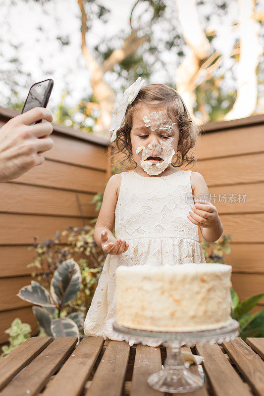 2019冠状病毒病(COVID-19)期间，在家中举办的简单生日派对上，刚学步的女儿撞到她的2岁生日蛋糕，父亲用手机拍照
