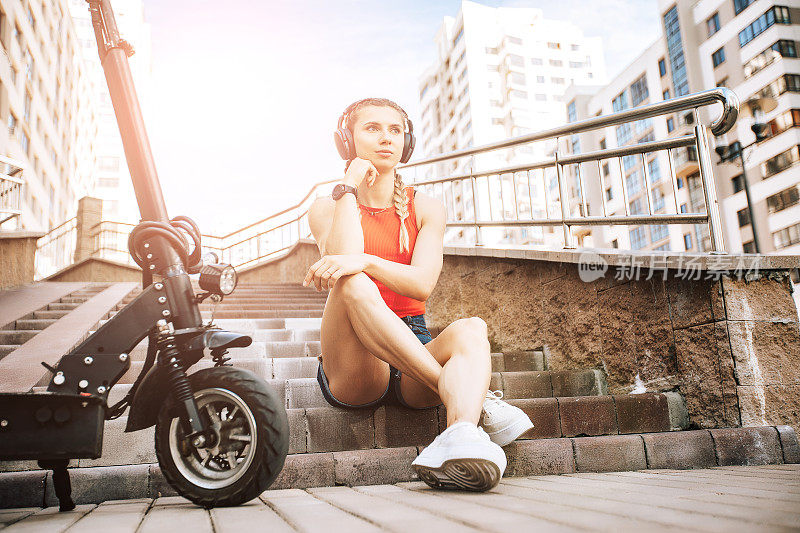 使用电动滑板车的女人的肖像。推滑板车概念户外街道。一个人骑着电动车坐在楼梯上放松。照片与音乐无线耳机