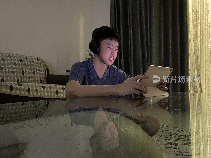 年轻的亚洲男孩戴着耳机在起居室用数字平板电脑玩视频游戏