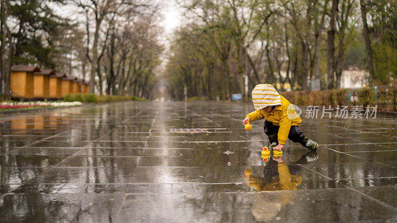 可爱活泼的小男孩穿着明黄色雨衣和胶靴在潮湿的春雨日街道上的小水坑里玩橡皮鸭。季节性天气步行概念