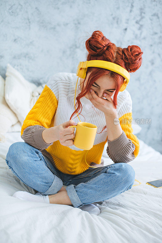 一个女孩坐在床上，喝着咖啡，笑着，戴着黄色的耳机听着音乐。