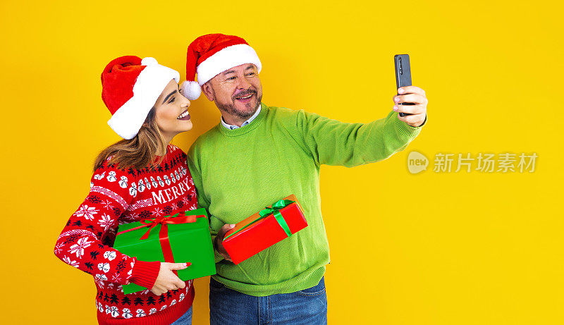 拉丁美洲墨西哥的父亲和女儿穿着红色毛衣，戴着圣诞帽，手里拿着圣诞礼物和gif，在黄色背景下自拍
