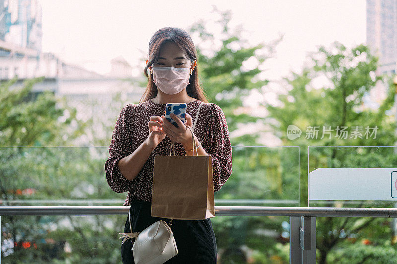 年轻的亚洲女性戴着口罩，手持购物袋，站在街上使用智能手机