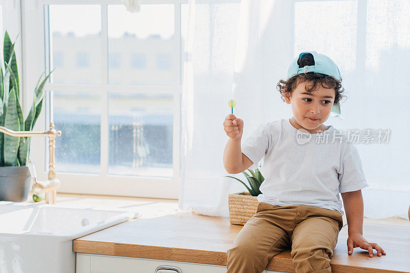 漂亮的卷曲的意大利小男孩，穿着白衬衫白t恤，棒球帽和裤子，拿着棒棒糖坐在厨房桌子上看着一边。英俊的西班牙男婴在家。童年,模型。