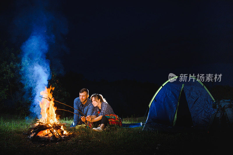一对夫妇在火上烤香肠，晚上在森林里的篝火旁放松。浪漫的篝火概念。