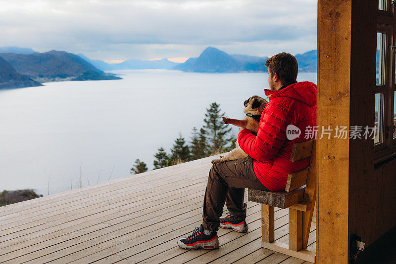 在挪威，一个快乐的男人带着狗在山上俯瞰风景优美的峡湾，沉思冬日