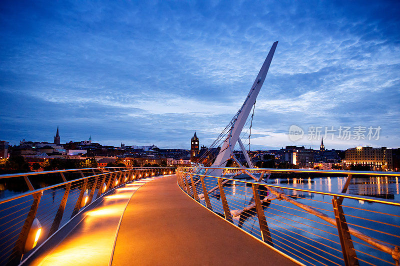 德里,爱尔兰。北爱尔兰文化之城伦敦德里的和平桥被灯光照亮，背景是市中心。夜晚多云的天空映着黄昏的河水