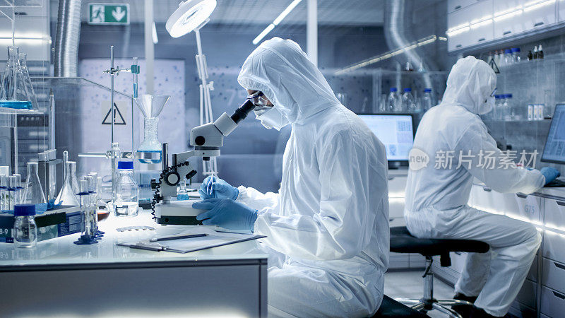在一个安全的高水平实验室里，科学家们穿着工作服进行一项研究。生物学家在有盖培养皿中用钳子调整样本并在显微镜下检查它们，他的同事在电脑上分析结果。