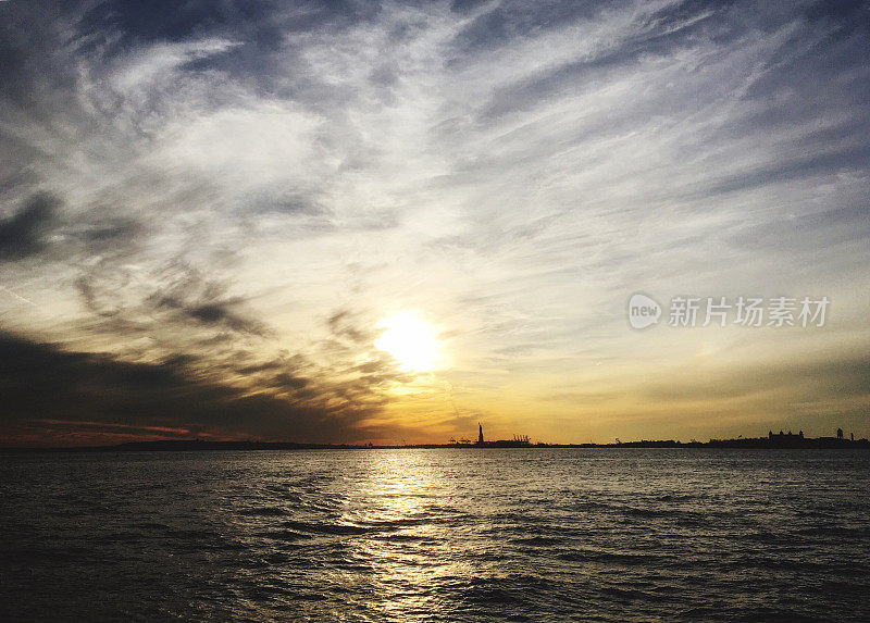 日落时分纽约港自由岛上的自由女神像。日落时戏剧性的天空和动态的云。远处的自由女神在日落时被水包围。
