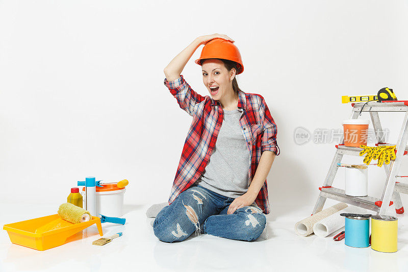 女人在橙色的保护头盔坐在地板上与仪器装修公寓房间隔离在白色的背景。墙纸，胶水配件，油漆工具。修理家里的概念。