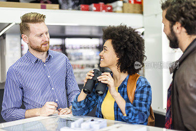 一个年轻的女人在一个相机和配件商店的售货员的帮助下选择一套双筒望远镜