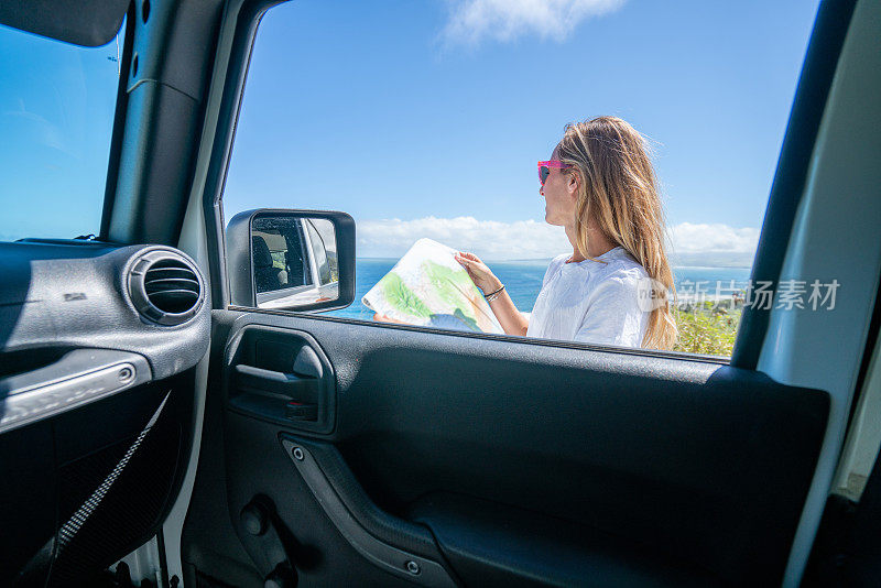 一名年轻女子在沿海公路上的车外看地图问路。夏威夷的山海景观