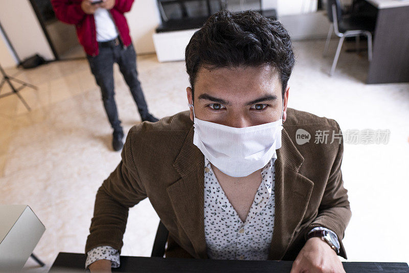 拉丁裔男子坐在办公室电脑前，戴着防护面具