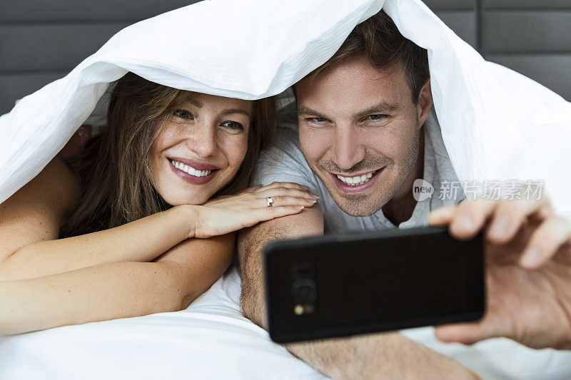 幸福浪漫的年轻夫妇躺在床上的羽绒被下自拍