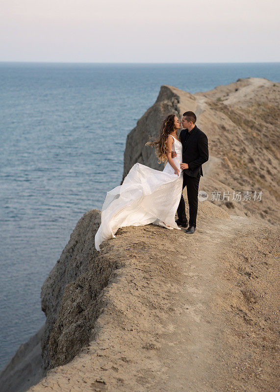 新郎和新娘在海景悬崖上拥抱。新娘的礼服在风中飘动。