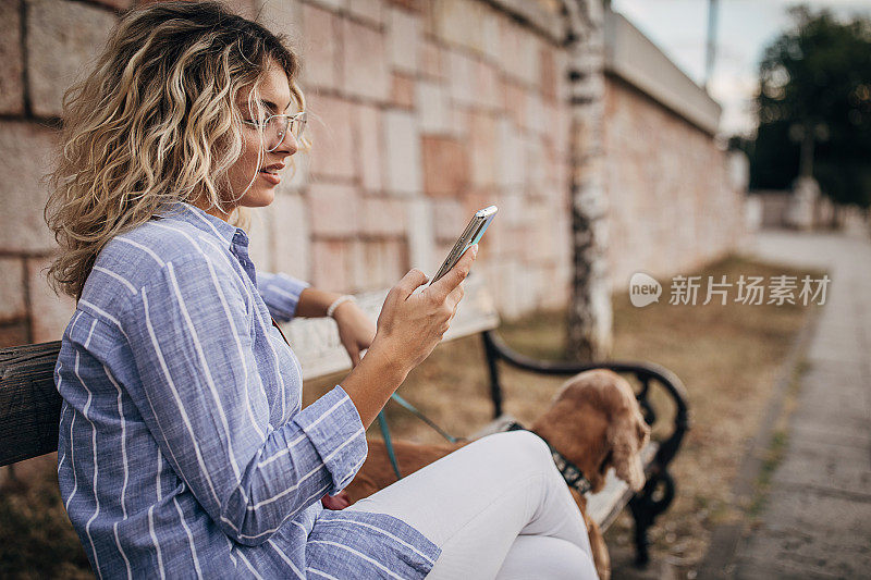 美丽的女人坐在长椅上与她的可可猎犬和使用智能手机