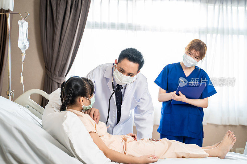 亚洲医生和护士戴着口罩在医院康复室检查年轻病人女孩的健康。感染新冠肺炎的孩子躺在床上接受医疗队的治疗和诊断。