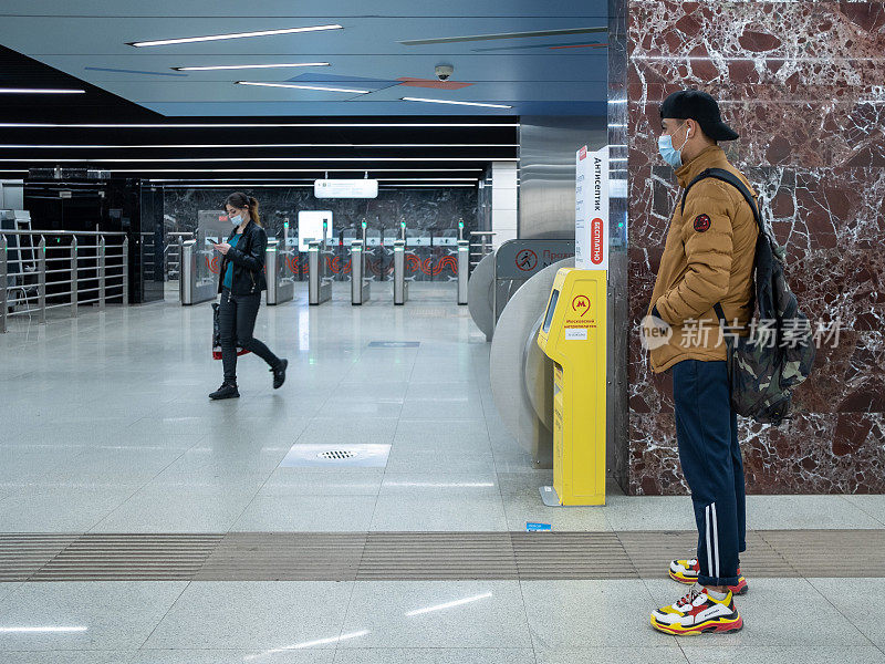一个戴着口罩的少年站在地铁站的大厅里。附近安装了专用的自动洗手液。