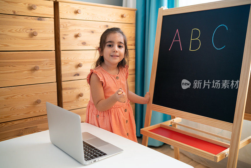 一个小女孩在家里学习字母，在黑板上写字，看着摄像机在线学习