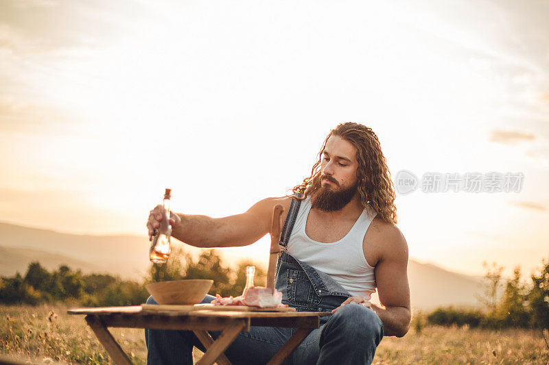 自然的守护者。一个在山上的男人，一个伐木工人，喝着自制的白兰地，吃着自制的食物，而在他身后是美丽的日落，休息辛苦的工作。