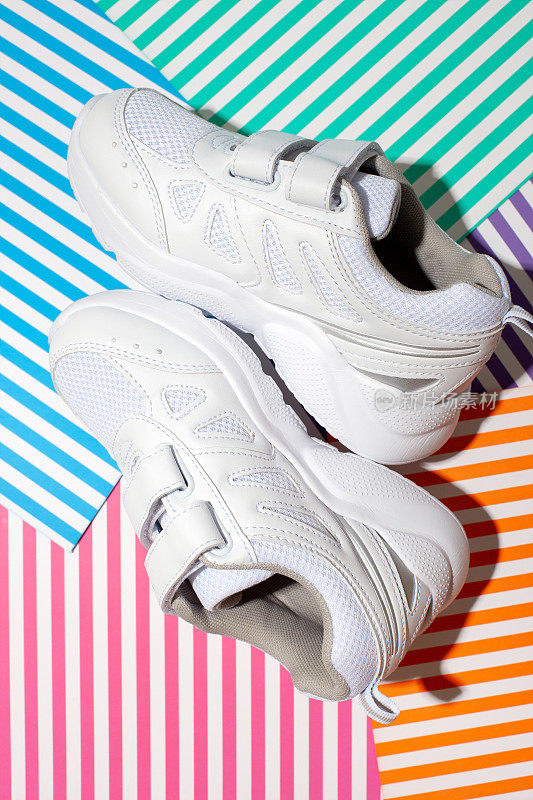 垂直俯视图两个白色的小孩运动鞋与尼龙搭扣为舒适的鞋在一个抽象的几何纸上彩色条纹背景与硬光