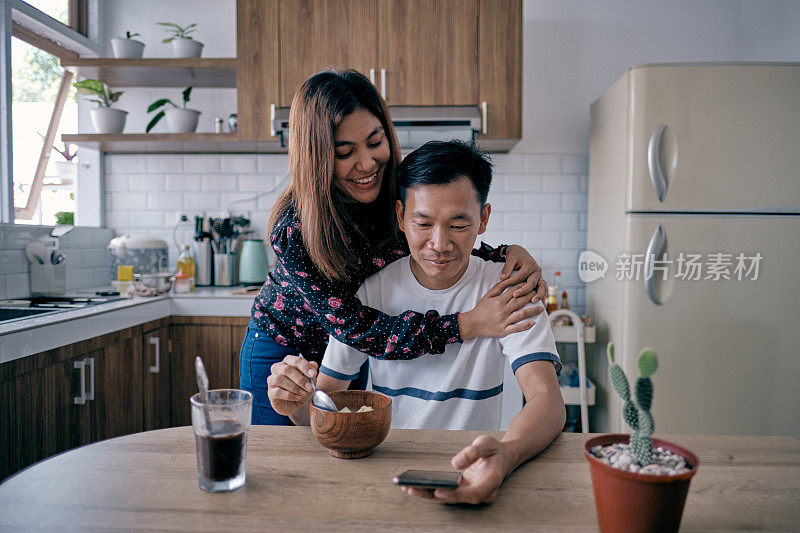 可爱的妻子在丈夫吃早餐时拥抱丈夫