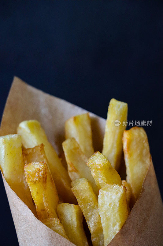 特写图像的防油羊皮纸包鲜煮酥脆的自制炸薯条，包裹堆金黄色的薯片零食，黑色背景，重点在前景