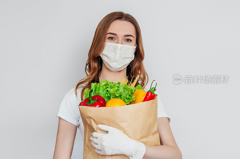 年轻的快递志愿者戴着医疗口罩，拿着一个纸袋，里面装着产品，蔬菜，辣椒，沙拉，白色，灰色的背景