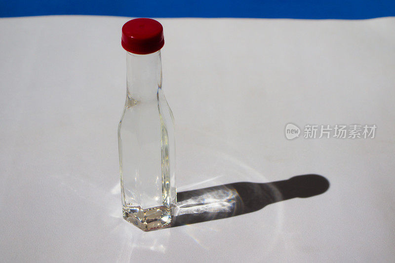 一个红色瓶盖的透明玻璃瓶的侧视图，里面装着无色的液体，在晨光的照射下，在白色的表面和蓝色的背景上投射出阴影和反射。