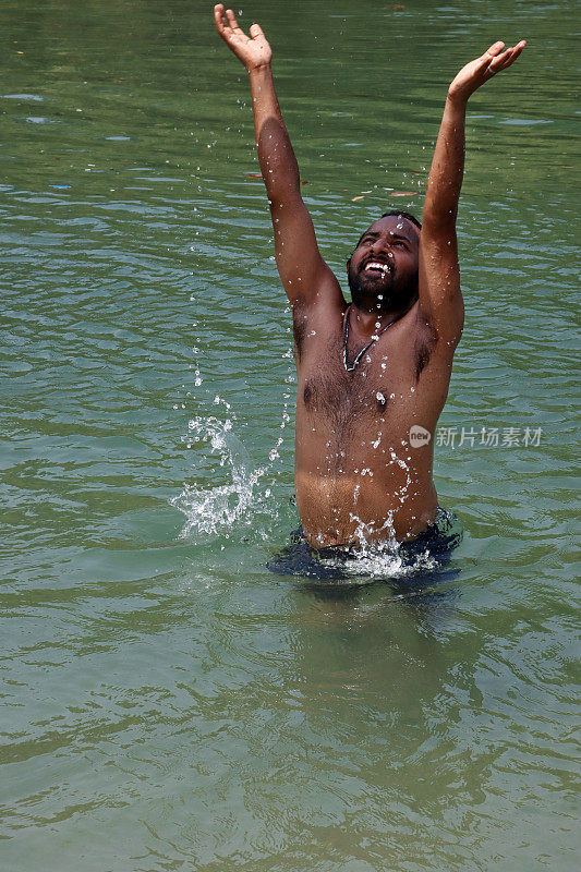 特写照片:一名印度男子穿着冲浪短裤站在齐腰深的泻湖平静的水中，笑着举起手臂，溅起水花，聚焦在前景上