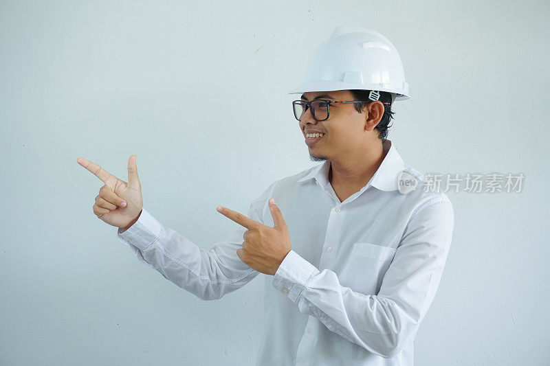 年轻的亚洲建筑师男子戴着白色安全帽，看着相机，手指指向右侧，孤立在白色背景上
